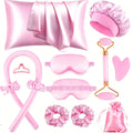 Collection of pink beauty and self-care products including a satin sheet, face mask, hair cap, brush, roller, and scrunchies on a white background.